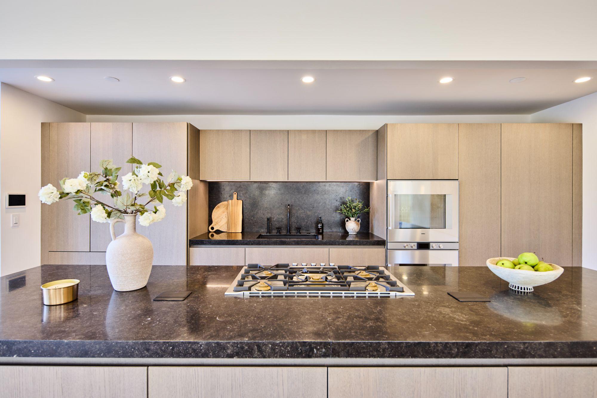 A modern kitchen with a gas hob set into a dark stone island and light wood cabinetry.
